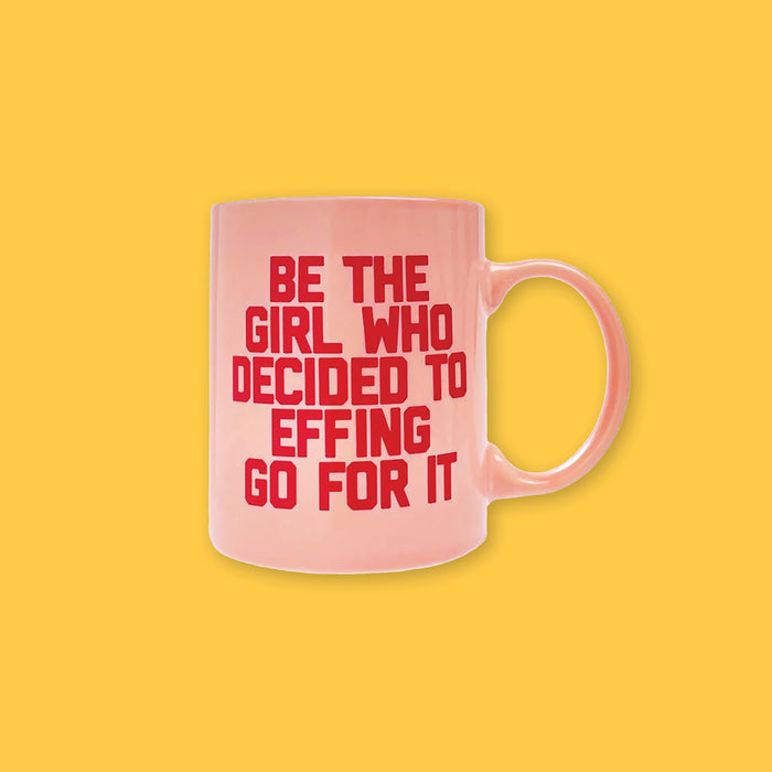 On a sunny mustard background sits a mug. This vintage inspired mug is light pink and has red wording on it. It says "BE THE GIRL WHO DECIDED TO EFFING GO FOR IT" in bold, block lettering.