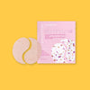 On a sunny mustard background sits a package with eye gels. The package is light pink and has a colorful illustration of two eye patches in white, purple, light pink, and gold. It says "patchology mood patch" in white font. It's called "HAPPY PLACE" and it says "inspiring tea-infused aromatherapy eye gels" in hot pink, serif font. It also says "ROSE HIBISCUS LOTUS FLOWER/FLEUR DE LOTUS" in hot pink, all caps block font. To the left is the two eye patches. 1 pair