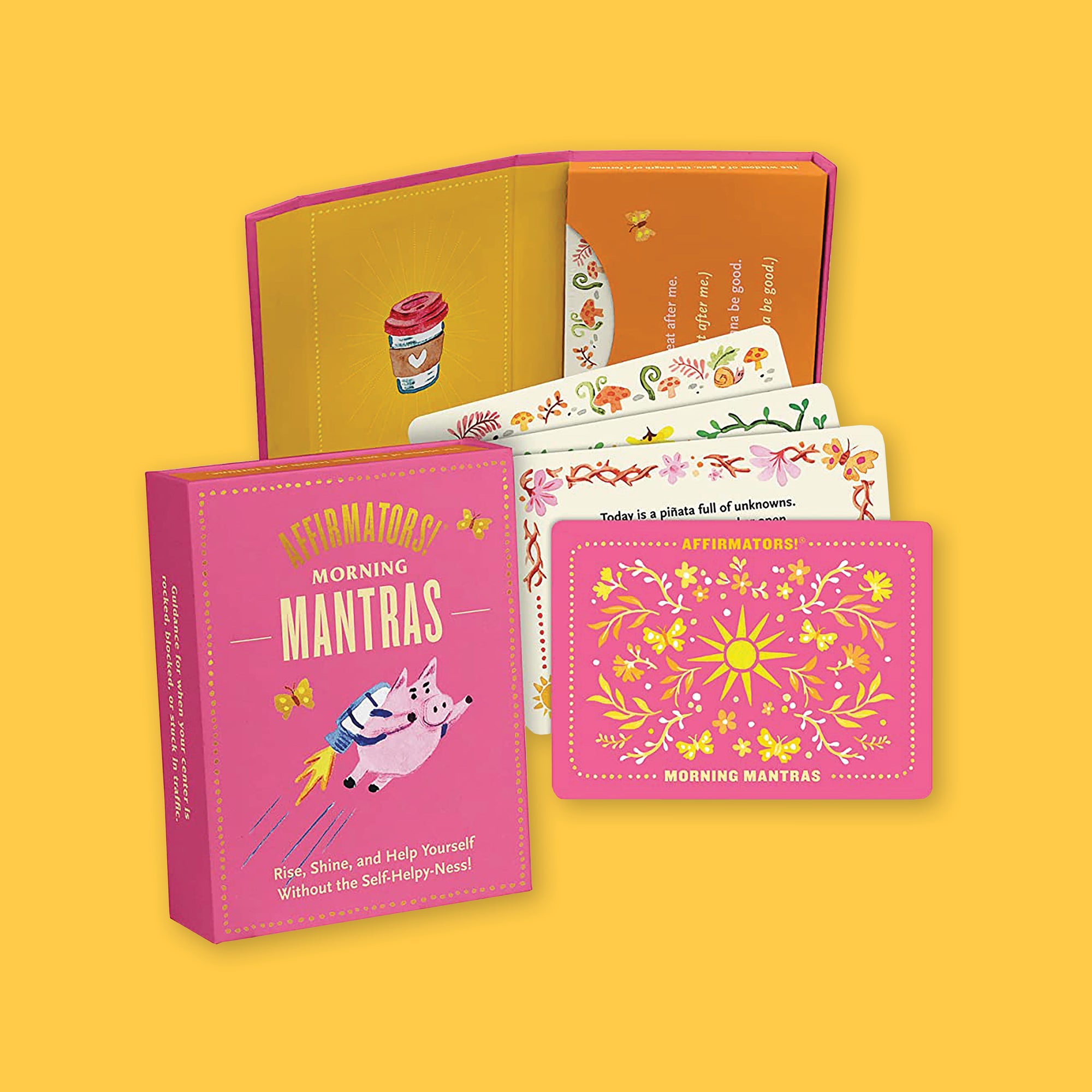 On a sunny mustard background sits a hot pink deck of cards box. It has an illustration of a pig with a rocket on it's back. It says "AFFIRMATORS! MORNING MANTRAS" in gold and yellow, all caps font. It also say "Rise, Shine, and Help Yourself Without the Self-Helpy-Ness!" in yellow font. To the right is an open case with cards in it and four cards laid out below it. The case has an illustration of a to-go coffee cup with a pink lid and a heart on the front. The cards have various colorful illustrations.