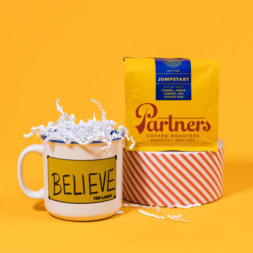 On a mustard yellow background sits a mug and a bag of coffee. The yellow bag of Partners coffee sits atop a roll of red and white striped tape. The mug is a white campfire style mug lined in blue with a yellow "Believe" sign from the hit show Ted Lasso. It's filled with white crinkle.