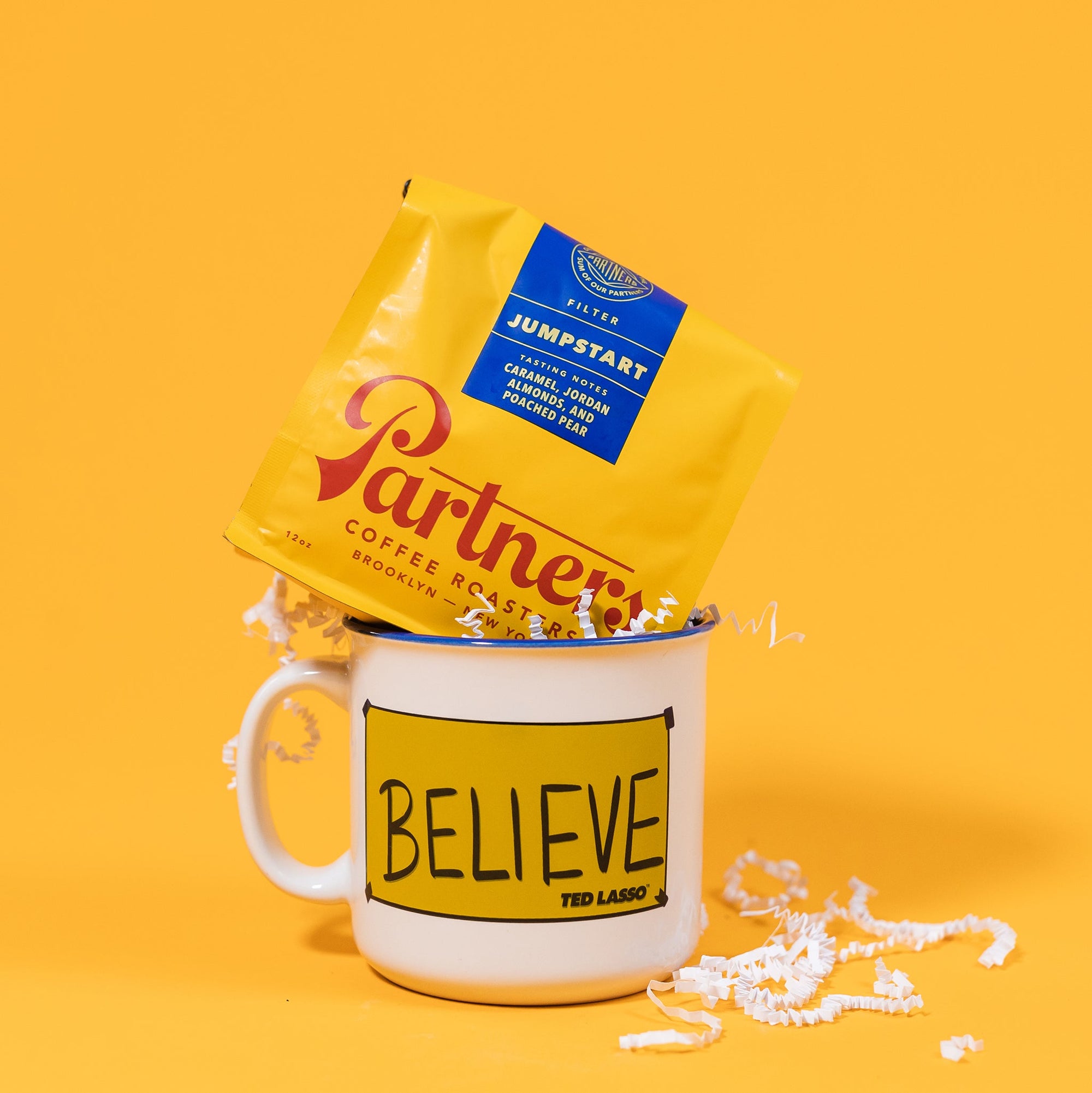 On a mustard yellow background sits a mug and a bag of coffee. The yellow bag of Partners coffee sits jauntily atop the mug. The mug is a white campfire style mug lined in Blue with a yellow "Believe" sign from the hit show Ted Lasso. It's filled with white crinkle.