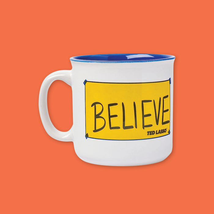 On an orangey-red background is a photo of a white campfire style mug with a blue interior. The mug is lined in Blue with a yellow "Believe" sign from the hit show Ted Lasso.
