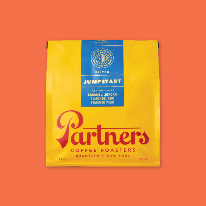 On an orangey-red background is a photo of the front of a yellow and blue bag of Partners Coffee Roasters Brooklyn - New York Jumpstart Blend. Region: Latin America; Tasting notes: Caramel, jordan almonds, and poached pear; 12 oz, 340 g