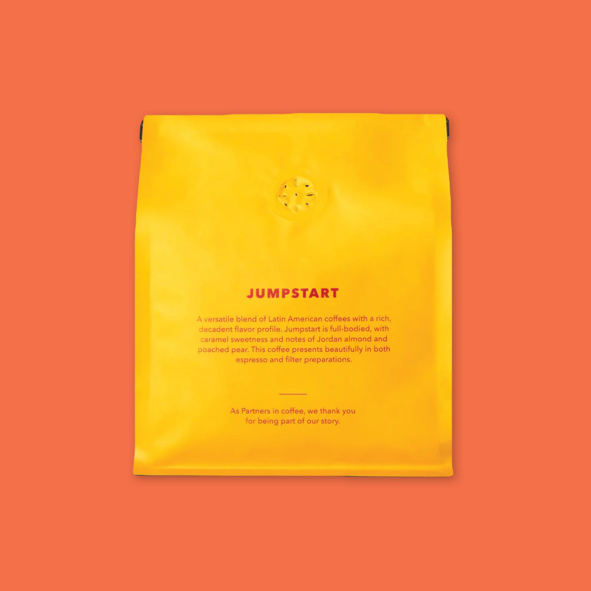 On an orangey-red background is a photo of the back of a yellow bag of Partners Coffee Roasters Jumpstart Blend. In red font reads "Jumpstart: A versitile blend of Latin American coffees with a rich, decadent flavor profile. Jumpstart is full-bodied, with caramel sweetness and notes of Jordan almond and poached pear. The coffee presents beautifully in both espresso and filtered preparations. As partners in coffee, we thank you for being part of our story."