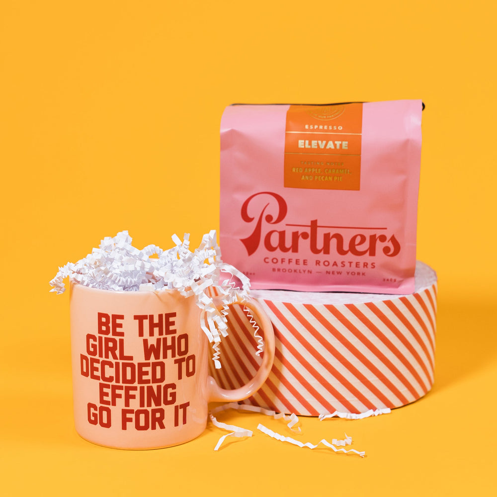 On a mustard yellow background sits a mug and a bag of coffee. The pink bag of Partners coffee sits atop a roll of red and white striped tape. The mug is a pink diner style mug with red capital collegiate writing that says "Be the girl who decided to effing go for it." White crinkle also overflows out of the mug.