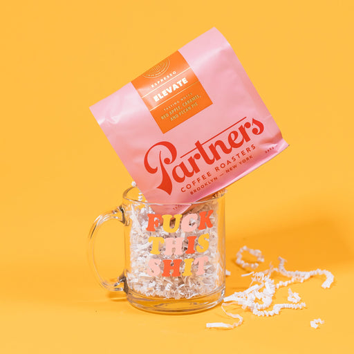 On a mustard yellow background sits a glass mug with bag of coffee sitting whimsical atop it. The clear glass mug says "FUCK THIS SHIT" in adorable red, yellow and pink cooper-esque font and is filled with white crinkle. The pink bag of Partners coffee is their "Elevate Blend"