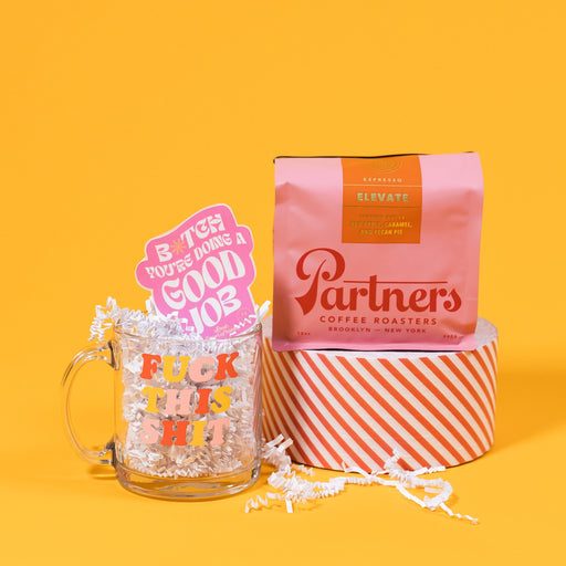 On a mustard yellow background sits a glass mug and bag of coffee. The clear glass mug says "FUCK THIS SHIT" in adorable red, yellow and pink cooper-esque font and is filled with white crinkle. The pink bag of Partners coffee sits atop a roll of red and white striped tape. 