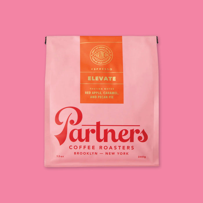 On a bubblegum pink background is a photo of a pink and red bag of Partners Coffee Roasters Brooklyn - New York Jumpstart Blend. Tasting notes: Red apple, caramel and pecan pie, 12 oz, 340 g