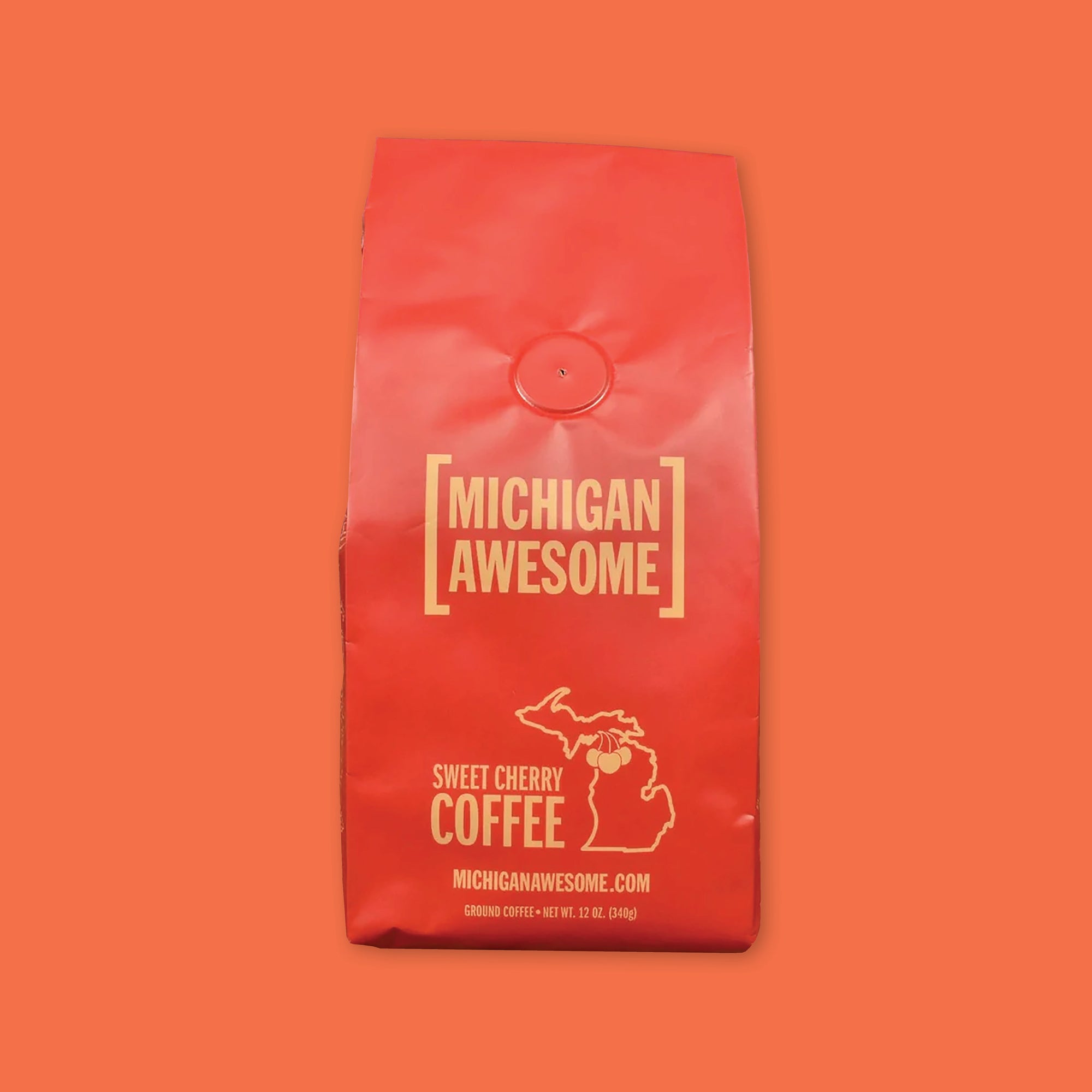 On an orangey red background is a red bag of coffee. The coffee is Michigan Awesome brand Sweet Cherry Ground Coffee. 12 oz (350 g)