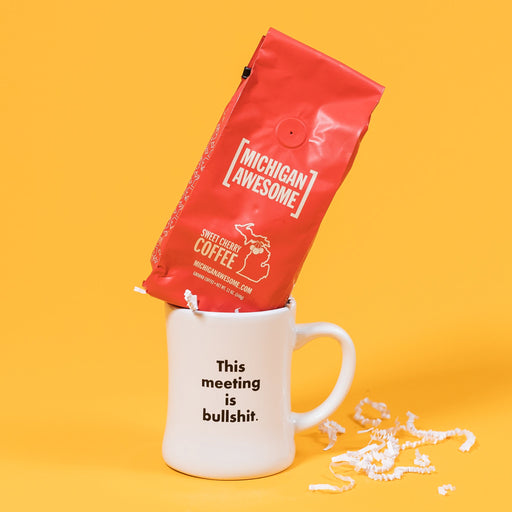On a mustard yellow background is a photo of a mug and a bag of coffee. The mug is a white diner style mug that says in black basic type, "This meeting is bullshit." A red bag of Michigan Awesome Sweet Cherry ground coffee sits atop the mug. White crinkle also overflows from the mug.