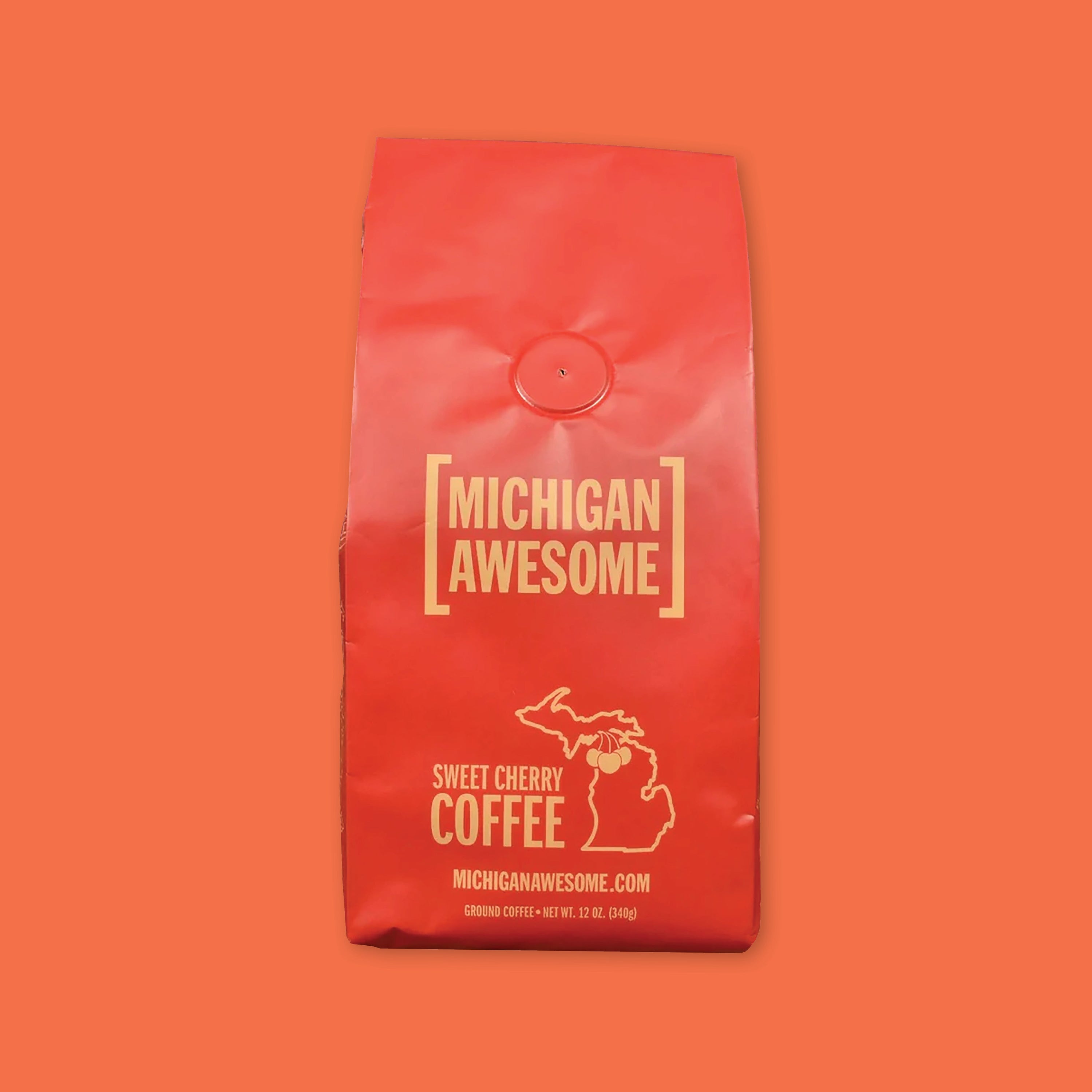 On an orangey red background is a red bag of coffee. The coffee is Michigan Awesome brand Sweet Cherry Ground Coffee. 12 oz (350 g)