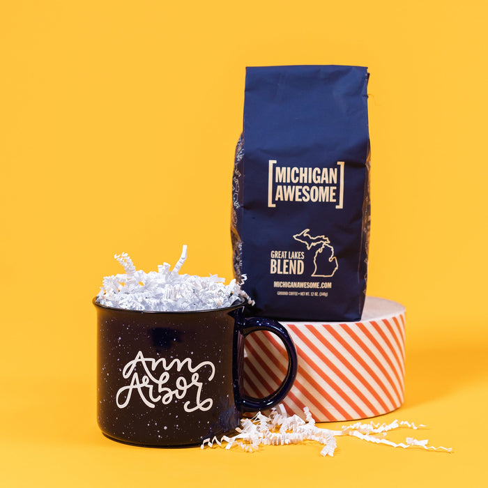 On a mustard yellow backdrop sits a mug and a bag of coffee. The mug is a navy blue campfire style mug with white splatter pattern. On the mug is white hand-lettering that says "Ann Arbor." The navy blue bag of coffee is Michigan Awesome brand Great Lakes Blend that sits atop a roll of red and white striped tape. Ground coffee. 12 oz (350 g)