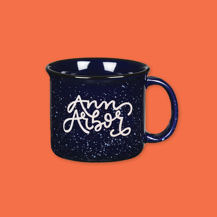 On an orangey-red backdrop is a navy blue diner style mug with a white splatter pattern. On the mug is white hand-lettering that says "Ann Arbor." 
