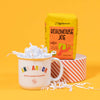 On a sunny mustard yellow and cool aqua backdrop sits a mug and a bag of coffee. The mug is a large white mug with colorful collegiate font that says "Ann Arbor" with "ROCK PAPER SCISSORS" below it. The bright yellow blue bag of coffee sits atop a red and white striped roll of tape. The coffee is Zingerman's brand Roadhouse Joe Blend. 'Smooth as silk and served daily at Zingerman's Roadhouse' Ground coffee. 12 oz (350 g). White crinkle overflows from the mug.