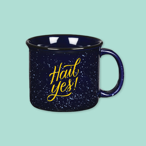 On a cool aqua backdroup is a navy blue campfire style mug with white splatter pattern. On the mug is a mustard yellow script font that says "Hail Yes!"