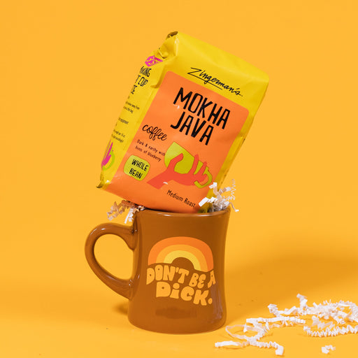 On a sunny mustard background sits a mug and a bag of coffee. The mug is a brown diner style mug with orange, groovy style font that says "Don't Be A Dick." with an orange, light orange and yellow rainbow above it. The Bright yellow orange bag of coffee sits atop the mug with white crinkle. The coffee is Zingerman's brand Roadhouse Joe Blend. 'Smooth as silk and served daily at Zingerman's Roadhouse' Ground coffee. 12 oz (350 g). White crinkle is scattered around.