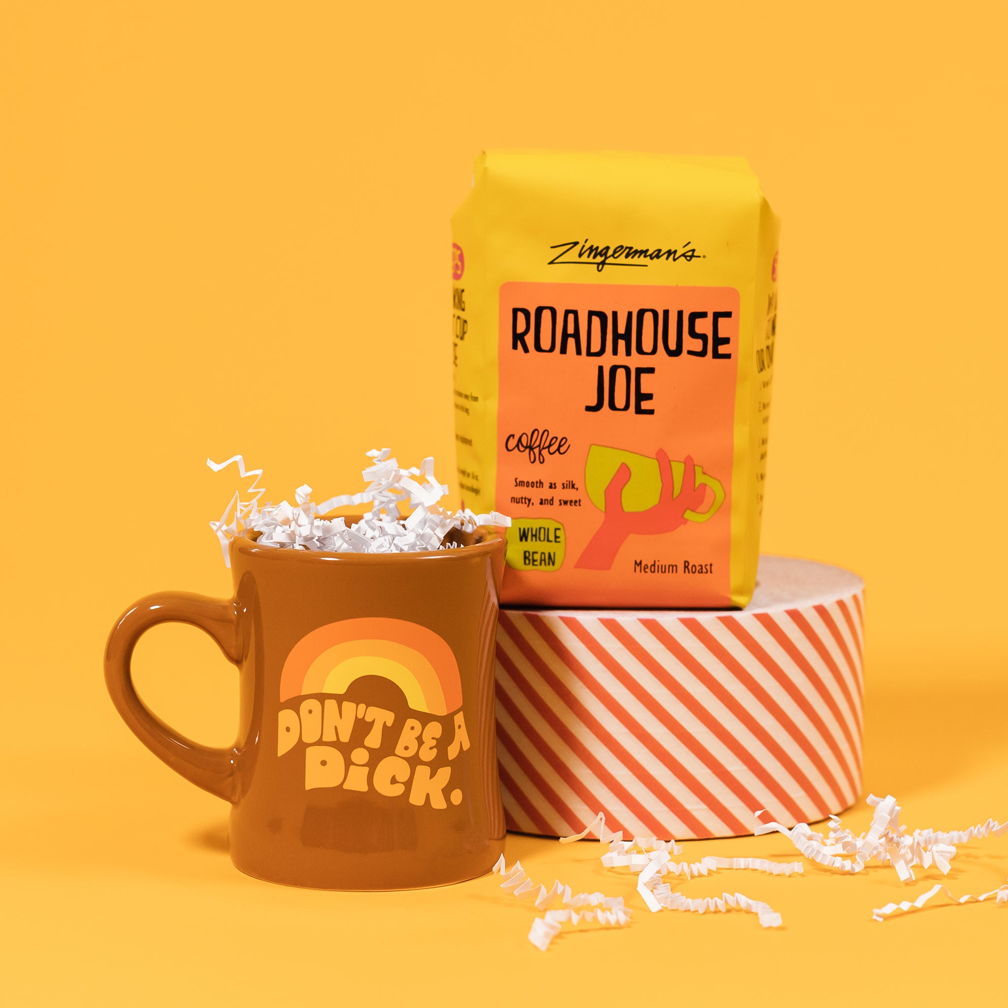 On a sunny mustard background sits a mug and a bag of coffee. The mug is a brown diner style mug with orange, groovy style font that says "Don't Be A Dick." with an orange, light orange and yellow rainbow above it. It has white crinkle in it. The bright yellow orange bag of coffee sits atop a red and white striped roll of tape. The coffee is Zingerman's brand Roadhouse Joe Blend. 'Smooth as silk and served daily at Zingerman's Roadhouse' Ground coffee. 12 oz (350 g). White crinkle is scattered around.