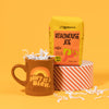On a sunny mustard background sits a mug and a bag of coffee. The mug is a brown diner style mug with orange, groovy style font that says "Don't Be A Dick." with an orange, light orange and yellow rainbow above it. It has white crinkle in it. The bright yellow orange bag of coffee sits atop a red and white striped roll of tape. The coffee is Zingerman's brand Roadhouse Joe Blend. 'Smooth as silk and served daily at Zingerman's Roadhouse' Ground coffee. 12 oz (350 g). White crinkle is scattered around.