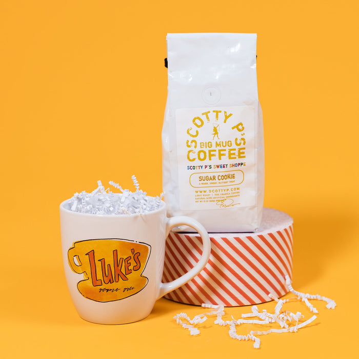 On a sunny mustard background sits a mug and a bag of coffee. The mug is a white, bistro-style mug with a Gilmore Girls-inspired "Luke's" watercolor illustration in red and yellows. It has white crinkle in it. The white bag of coffee sits atop a red and white striped roll of tape. The coffee is Scotty P's Coffe Sugar Cookie Blend. It is a "warm, sweet, buttery treat." 12 oz (350g). There is white crinkle scattered around.