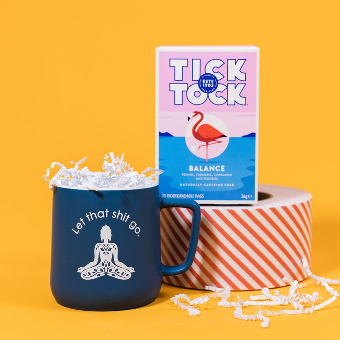 On a sunny mustard background sits a blue mug with white on the inside and on the brim. There is a buddha meditation pose in white, with white block lettering that says "Let that shit go." It is filled with white crinkle. The box of tea sits atop a red and white striped roll of tape. The tea is Tick Tock Balance with blends of fennel, turmeric, cinnamon and rooibos and it is naturally caffeine free. There is an illustration of a flamingo standing on one leg. There is white crinkle scattered around. 
