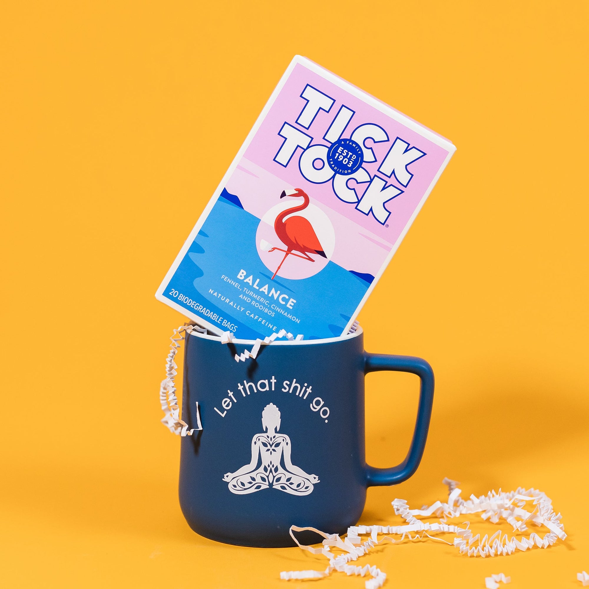 On a sunny mustard background sits a blue mug with white on the inside and on the brim. There is a buddha meditation pose in white, with white block lettering that says "Let that shit go." It is filled with white crinkle. The box of tea sits atop the mug. The tea is Tick Tock Balance with blends of fennel, turmeric, cinnamon and rooibos and it is naturally caffeine free. There is an illustration of a flamingo standing on one leg. There is white crinkle scattered around. 