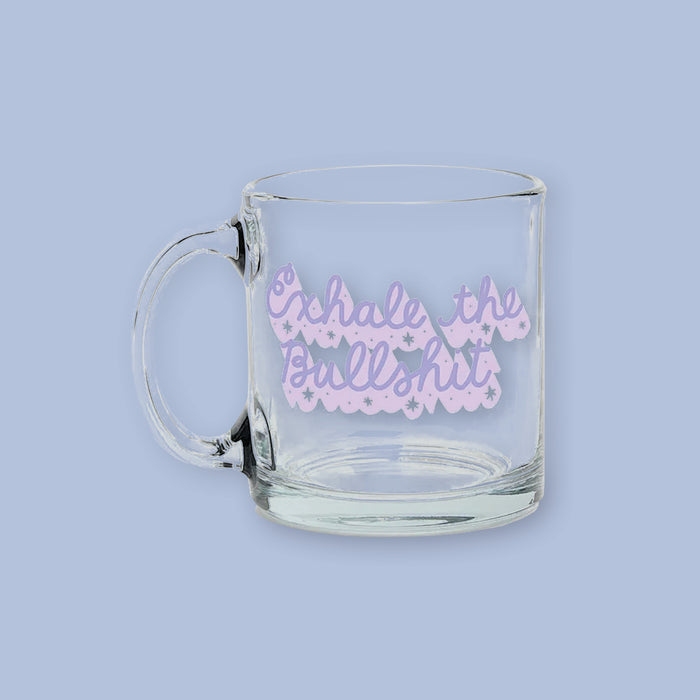 On a lavender background sits a clear mug with handwritten font that says "Exhale the Bullshit" in lavender and has a white dropshadow with stars. 