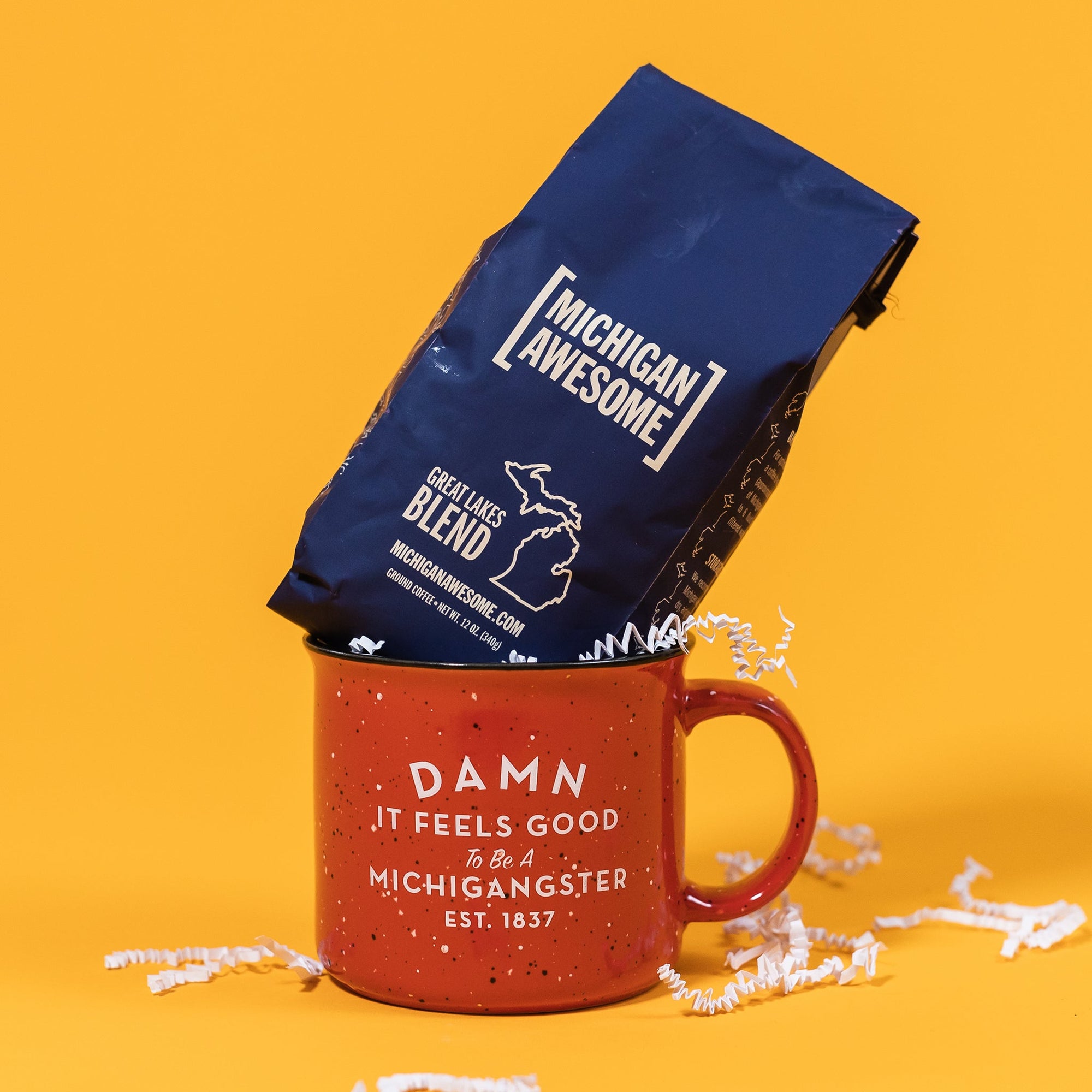 On a sunny mustard background sits a mug and a bag of coffee. The mug is a red campfire style mug with black and white splatter pattern. On the mug is a white block and script font that says "Damn It Feels Good To Be A Michigangster" with "EST. 1837" on the bottom. There is a navy bag of coffee sitting atop the mug. This Michigan Awesome Coffee is a Great Lakes Blend. 12 oz (350g). There is white crinkle scattered around.