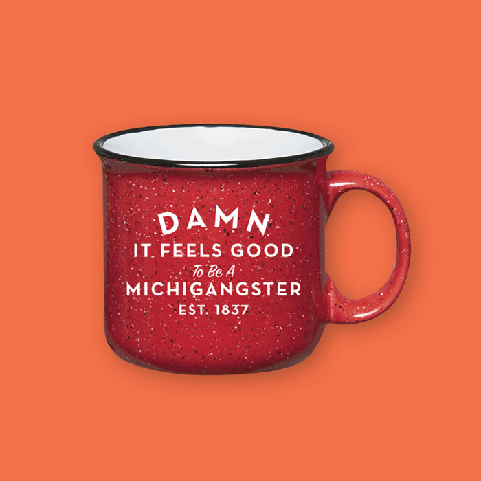 On a coral orange background sits a red campfire style mug with black and white splatter pattern. On the mug is a white block and script font that says "Damn It Feels Good To Be A Michigangster" with "EST. 1837" on the bottom.