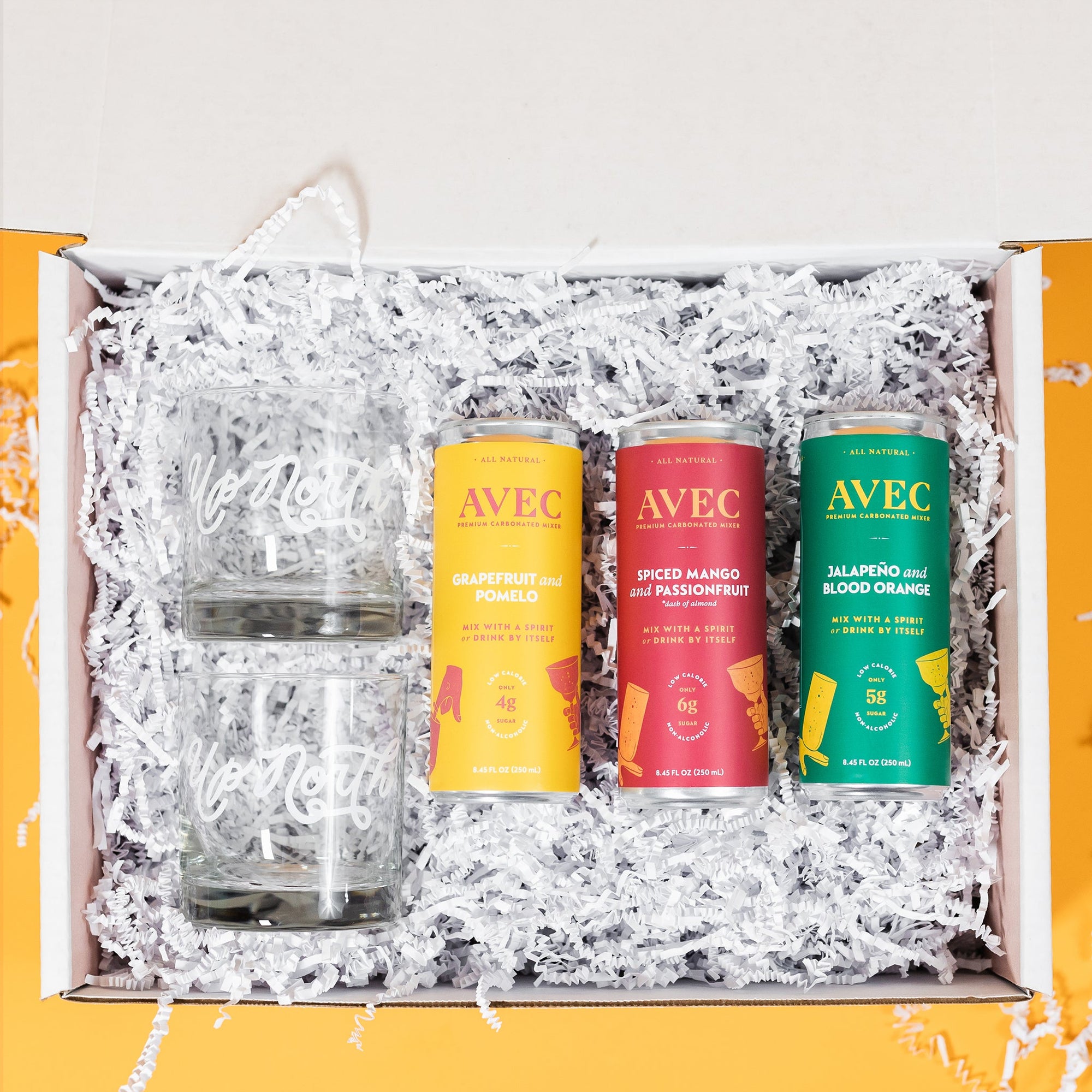 On a sunny mustard background sits a box with two high ball glasses and three cans of carbonated mixers with white crinkle underneath. The two glasses are clear with "Up North" handwritten in white. There are three cans of AVEC premium carbonated mixers. The mustard yellow can is Grapefruit and Pomelo (4g sugar). The fuchsia can is Spiced Mango and Passionfruit (6g sugar) and the green can is Jalapeno and Blood Orange (5g sugar). 
