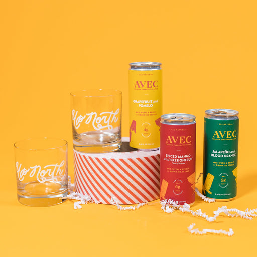 On a sunny mustard background sits two high ball glasses that are clear with "Up North" handwritten in white. There are three cans of AVEC premium carbonated mixers. The mustard yellow can is Grapefruit and Pomelo (4g sugar). The fuchsia can is Spiced Mango and Passionfruit (6g sugar) and the green can is Jalapeno and Blood Orange (5g sugar). One highball glass and the AVEC Grapefruit and Pomelo can is atop a red and white striped packing tape with white crinkle scattered around.