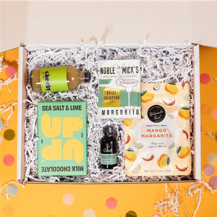 On a sunny mustard background sits a box filled with margarita goodies and white crinkle. There are big, colorful confetti surrounding the box. The UP UP Sea Salt & Lime Chocolate bar has lime green and yellow packaging, Noble Mick's Spicy Jalapeno Margarita Mix, Mango Margarita Dried Fruit Jerky, Cheeky Lime Juice and W&P Spicy Margarita Craft Cocktail Syrup. 