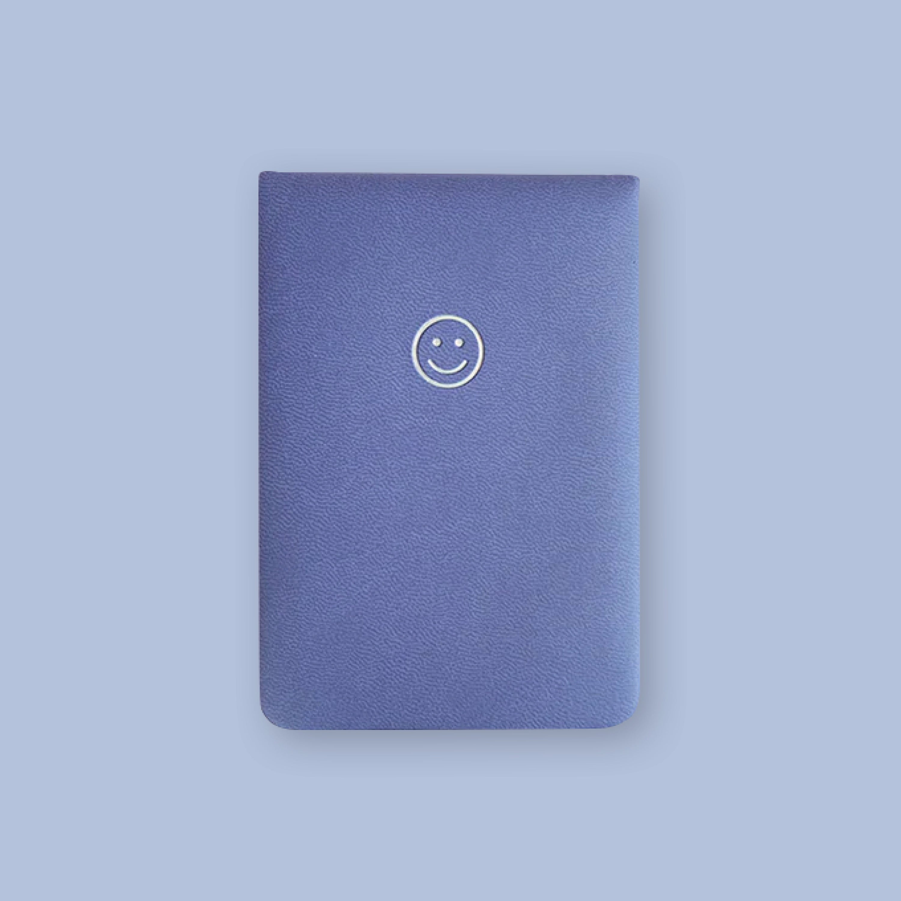 On a lavender background sits a rich, purple leather notebook with a smiley face on the top. 