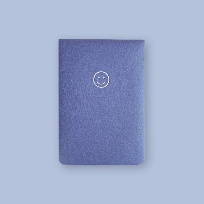 On a lavender background sits a rich, purple leather notebook with a smiley face on the top. 