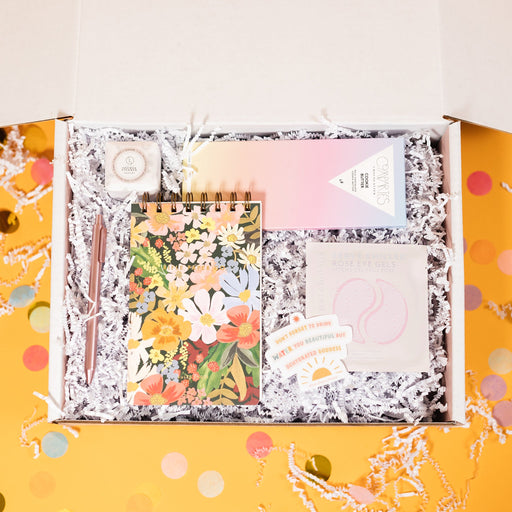 On a sunny mustard background sits a box filled with pretty pastel gifts on a bed of white crinkle. The gifts include a floral spiral notepad from Rifle Paper Co., Compartes Milk Chocolate Cookie Butter, RPS Exclusive Sticker that says "Don't Forget to Drink Water You Beautiful But Dehydrated Goddess" in a handwritten block, font with a yellow sun and the words "Rock Paper Scissors" in a handwritten script font under it, Lizush Lavender Shower Steamer, and Patchology Happy Place Eye Gel Patches. 