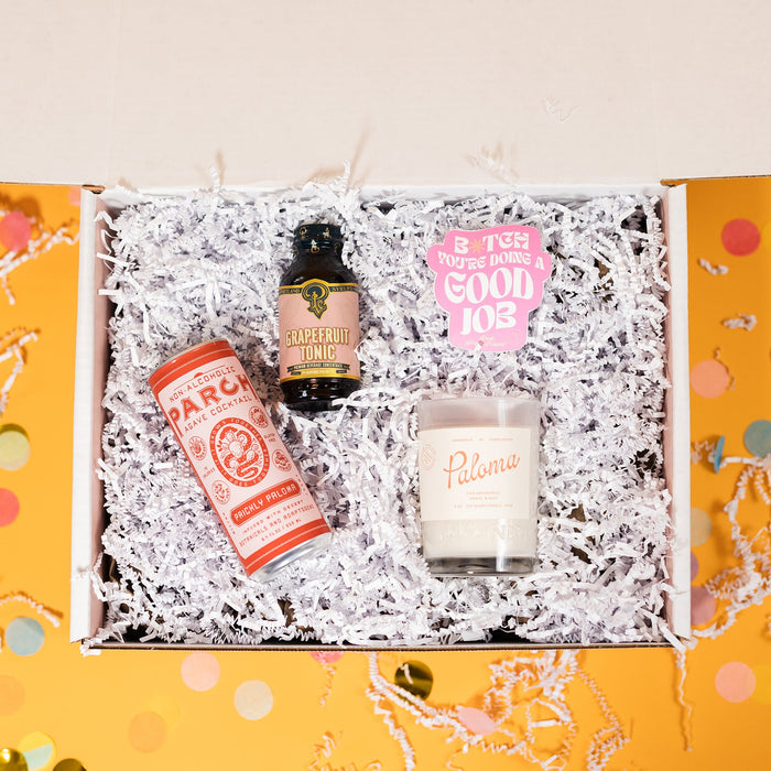 On a sunny mustard background sits a box filled with goodies for a Paloma Party, on a bed of white crinkle and big, round confetti scattered around. The goodies include Parch Non-Alcoholic Prickly Paloma Mix, Portand Syrups Grapefruit Tonic, handmade soy blend Paloma candle, and an RPS Exclusive purple sticker that says "B*tch You're Doing A Good Job" in a white, groovy font with "Rock Paper Scissors" handwritten below it in yellow with a smiley face. 