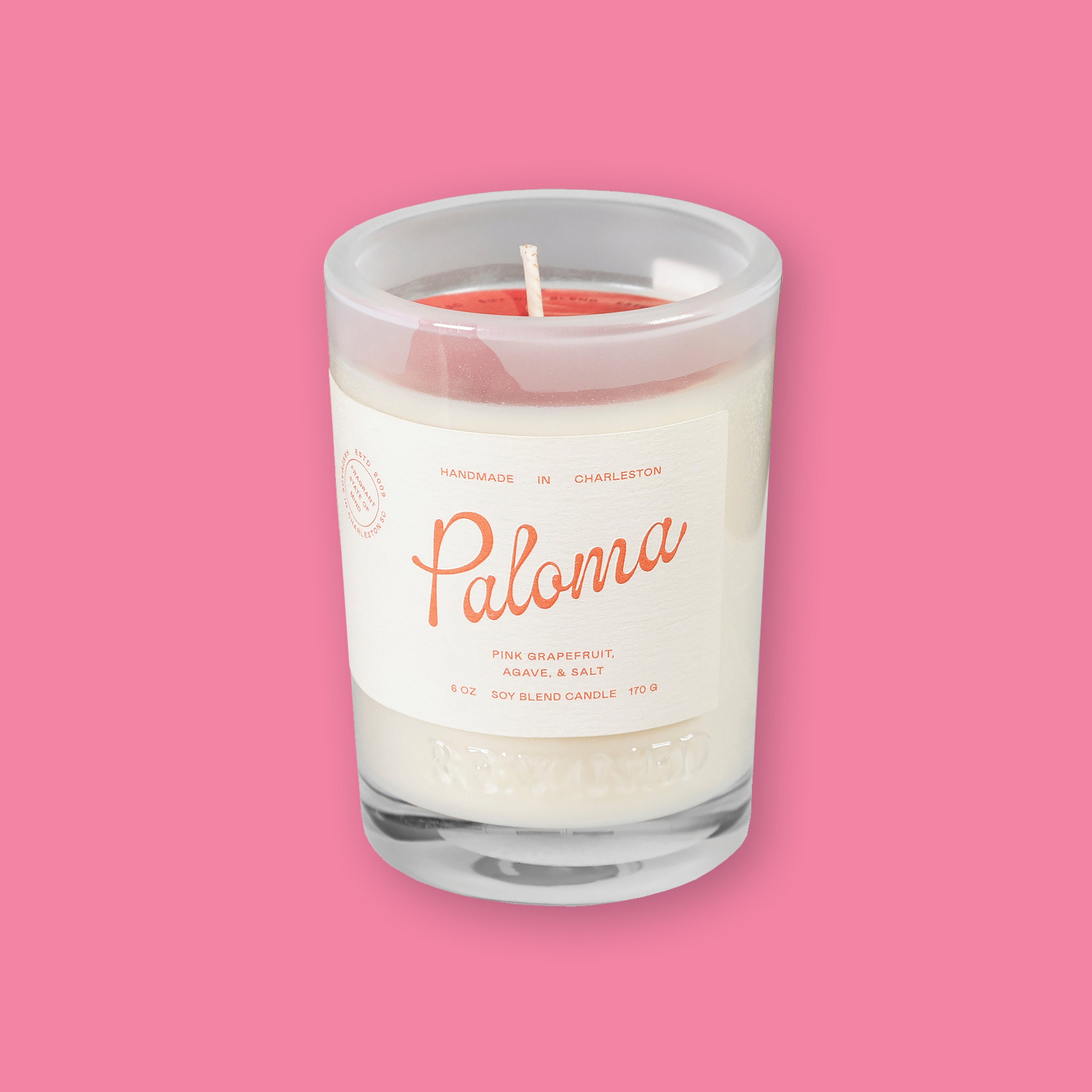 On a bubblegum pink background sits a Rewined Sparkling Collection Handmade Paloma Soy Blend Candle. It's scent is pink grapefruit, agave, and salt. It has a cream label with coral text.