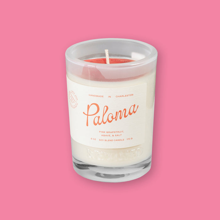 On a bubblegum pink background sits a Rewined Sparkling Collection Handmade Paloma Soy Blend Candle. It's scent is pink grapefruit, agave, and salt. It has a cream label with coral text.