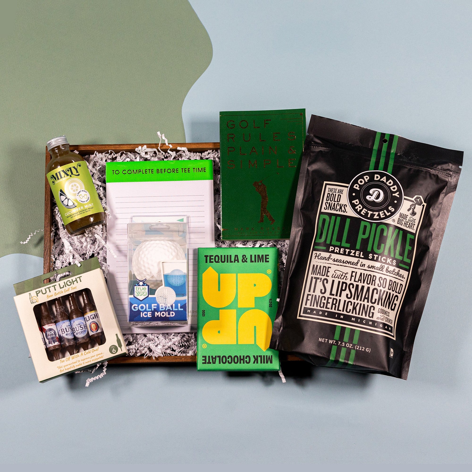 On a blue and green background sits a collection of items on white crinkle paper for a Father's Day gift box. A box of Putt Light beer bottle golf tees, and Golf Ball Ice Mold, a Tequila and Lime Milk Chocolate Bar, a black bag of Dill Pickle Pretzel Sticks, a green book titled "Golf Rules Made Simple", and notepad that says "To Complete Before Tee Time" and a small glass bottle of green Cucumber Mint Lime cocktail mix.