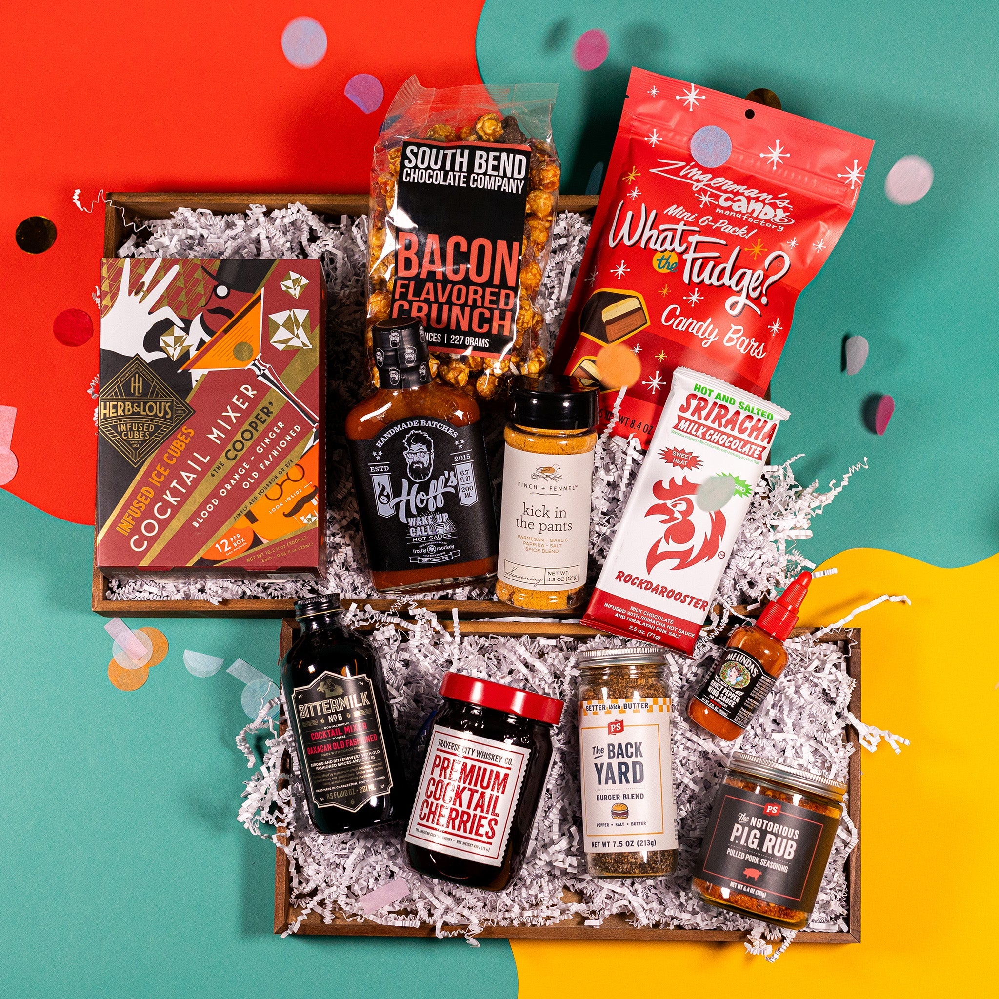 On an orange, teal, and yellow background with colorful confetti sits a collection of items in a gift box with white crinkle paper. A box of Infused Ice Cubes, a bag of Bacon Flavored Crunch popcorn, a bag of What the Fudge Candy Bars, a Sriracha flavored chocolate bar, a jar of Kick in the Pants seasoning, a bottle of Hoff's Wakeup Call hot sauce, a bottle of Bittermilk Cocktail Mixer, a jar of Premium Cocktail Cherries, a jar of Burger Blend seasoning, a bottle of wing sauce, and a jar of pork seasoning.