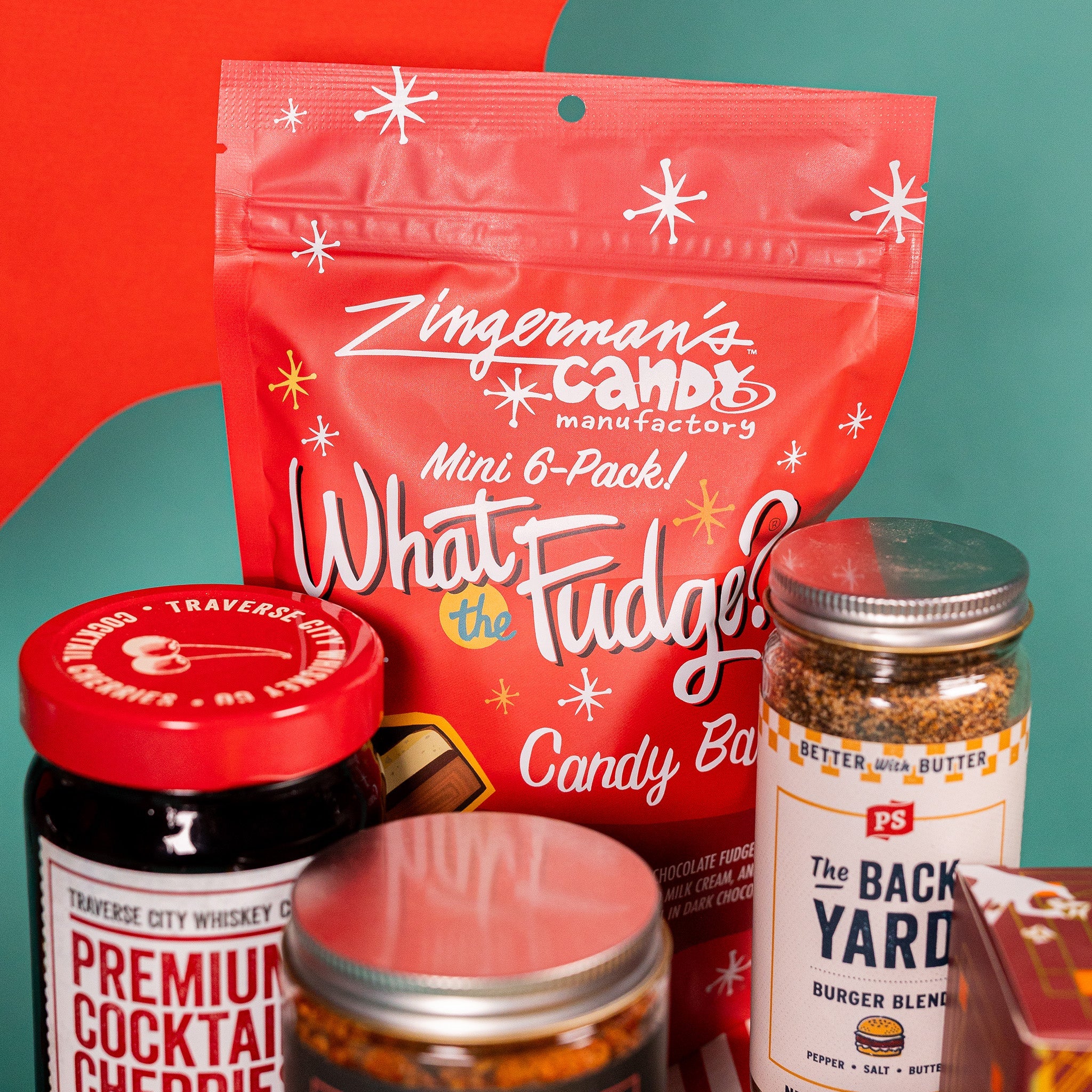 On a red and teal background sits a red bag of Zingerman's candy bars, a jar of The Back Yard Burger Blend, and a jar of Premium Cocktail Cherries with a red lid.