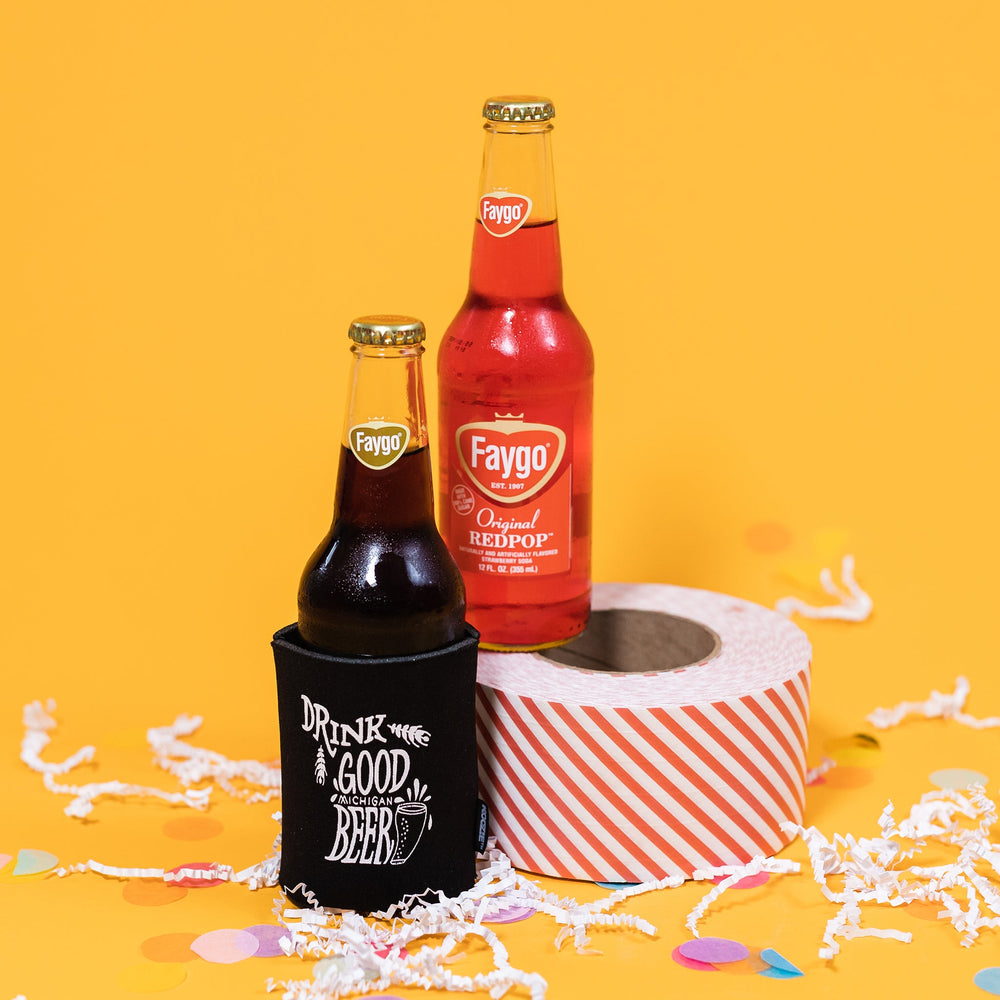 On a sunny mustard background sits a koozie and two bottles of pop with white crinkle and big, colorful confetti scattered around. The Faygo Original Root Beer is inside of a black koozie with white handdrawn illustration and text that says "Drink Good Michigan Beer". The Faygo Original Redpop is sitting atop a red and white striped packing tape. 12 oz (355ml)