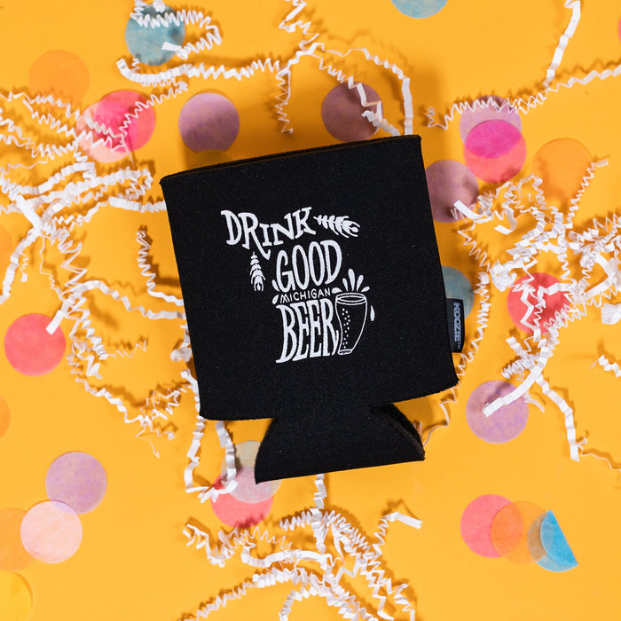 On a sunny mustard background sits a koozie with white crinkle and big, colorful confetti scattered around. The black koozie has white handdrawn illustration and text that says "Drink Good Michigan Beer."
