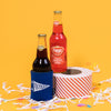 On a sunny mustard background sits a koozie and two bottles of pop with white crinkle and big, colorful confetti scattered around. The Faygo Original Root Beer is inside of a blue koozie with white handdrawn illustration of a pennant with handdrawn script font that says "Michigan." The Faygo Original Redpop is sitting atop a red and white striped packing tape. 12 oz (355ml)