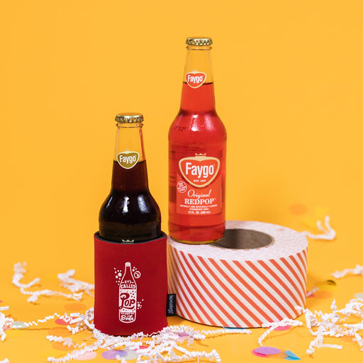 On a sunny mustard background sits a koozie and two bottles of pop with white crinkle and big, colorful confetti scattered around. The Faygo Original Root Beer is inside of a red koozie with white handdrawn illustration of a pop with handdrawn lettering that says "It's Called Pop Not Soda." The Faygo Original Redpop is sitting atop a red and white striped packing tape. 12 oz (355ml)