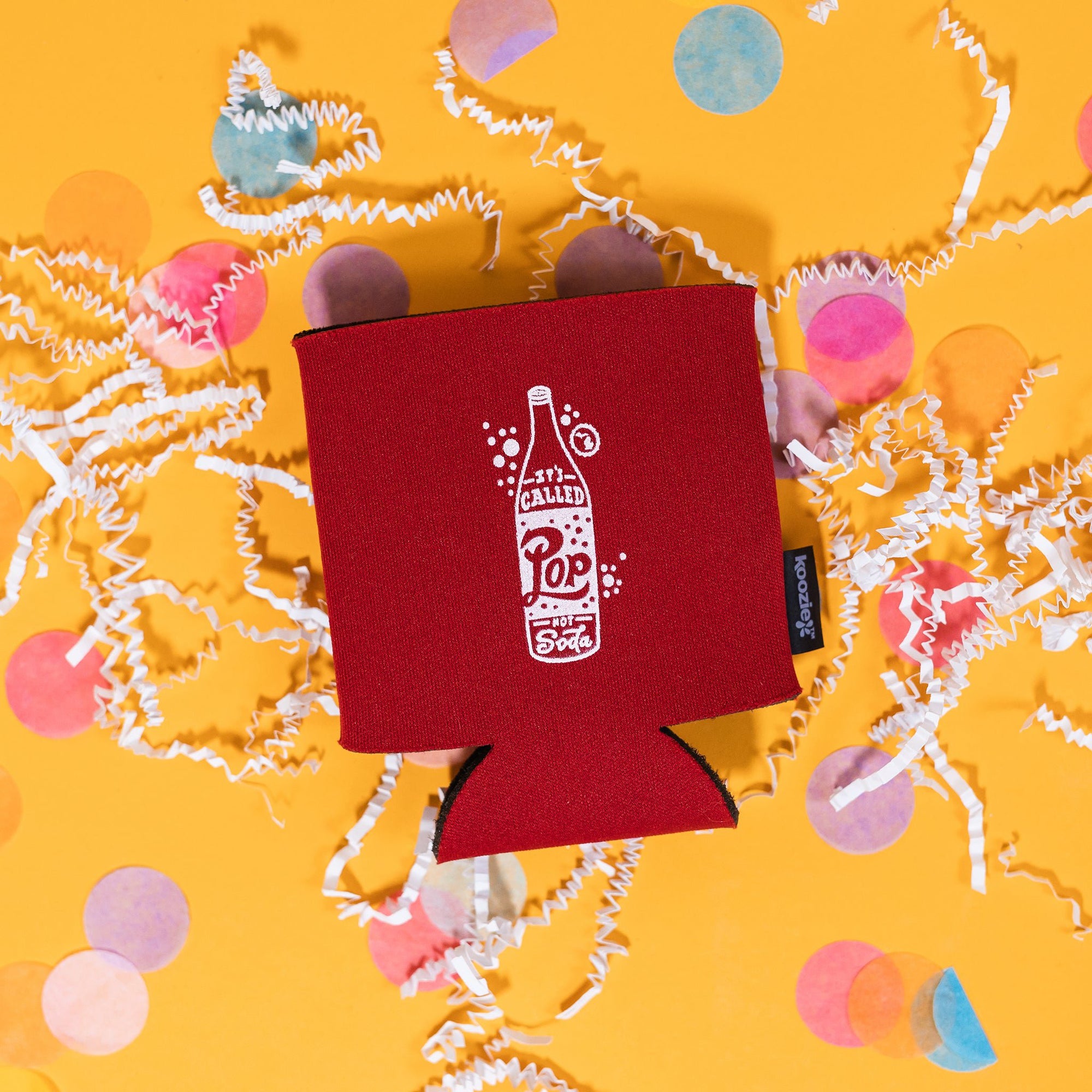 On a sunny mustard background sits a koozie with white crinkle and big, colorful confetti scattered around. The red koozie has a white handdrawn illustration of a pop with handdrawn lettering that says "It's Called Pop Not Soda."