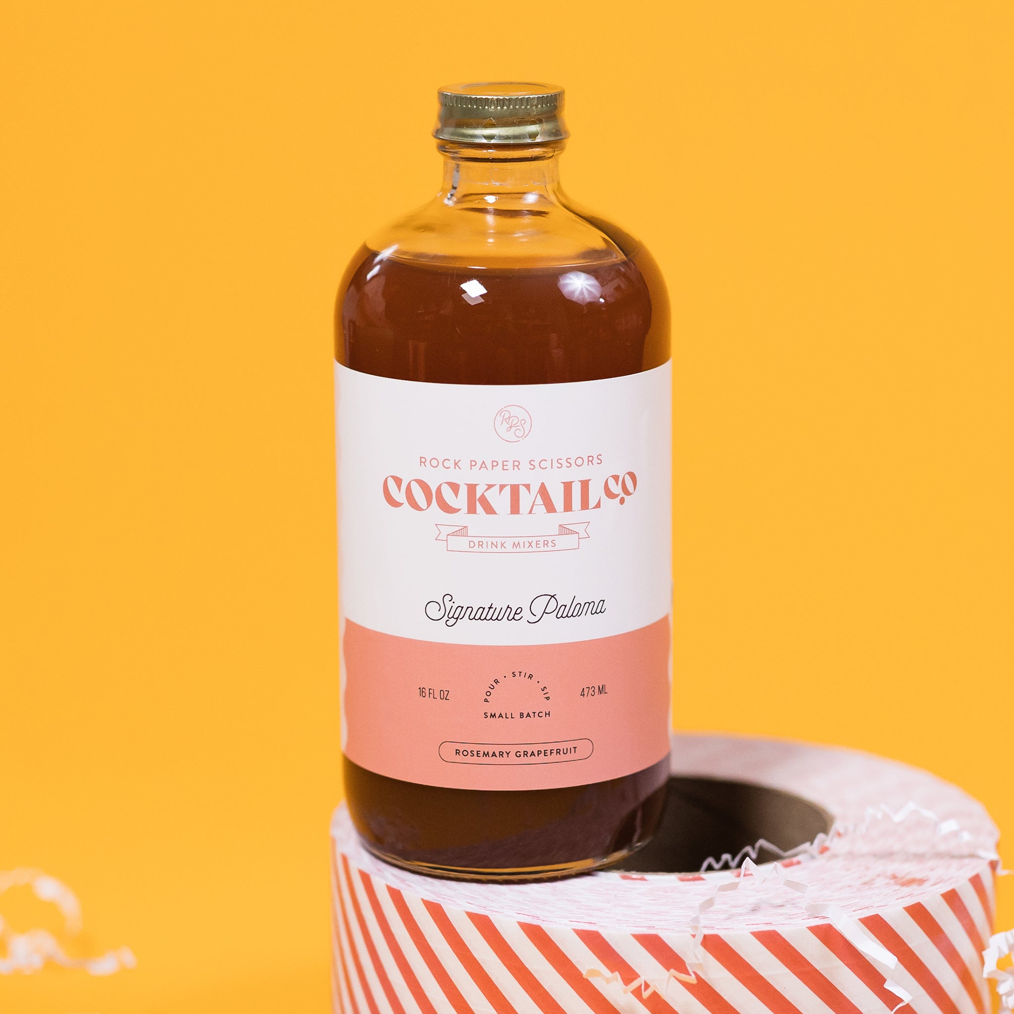 On a sunny mustard background sits a bottle and packing tape surrounded by white crinkle. The clear bottle is a Rock Paper Scissors Cocktail Co. Drink Mixer called "Signature Paloma". The flavor is rosemary grapefruit. It has a white label with a grapefruit pink stripe at the bottom. The bottle sits atop a red and white striped packing tape. 16 oz (473ml)