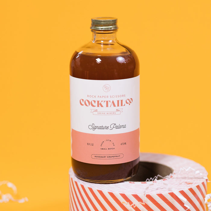 On a sunny mustard background sits a bottle and packing tape surrounded by white crinkle. The clear bottle is a Rock Paper Scissors Cocktail Co. Drink Mixer called "Signature Paloma". The flavor is rosemary grapefruit. It has a white label with a grapefruit pink stripe at the bottom. The bottle sits atop a red and white striped packing tape. 16 oz (473ml)