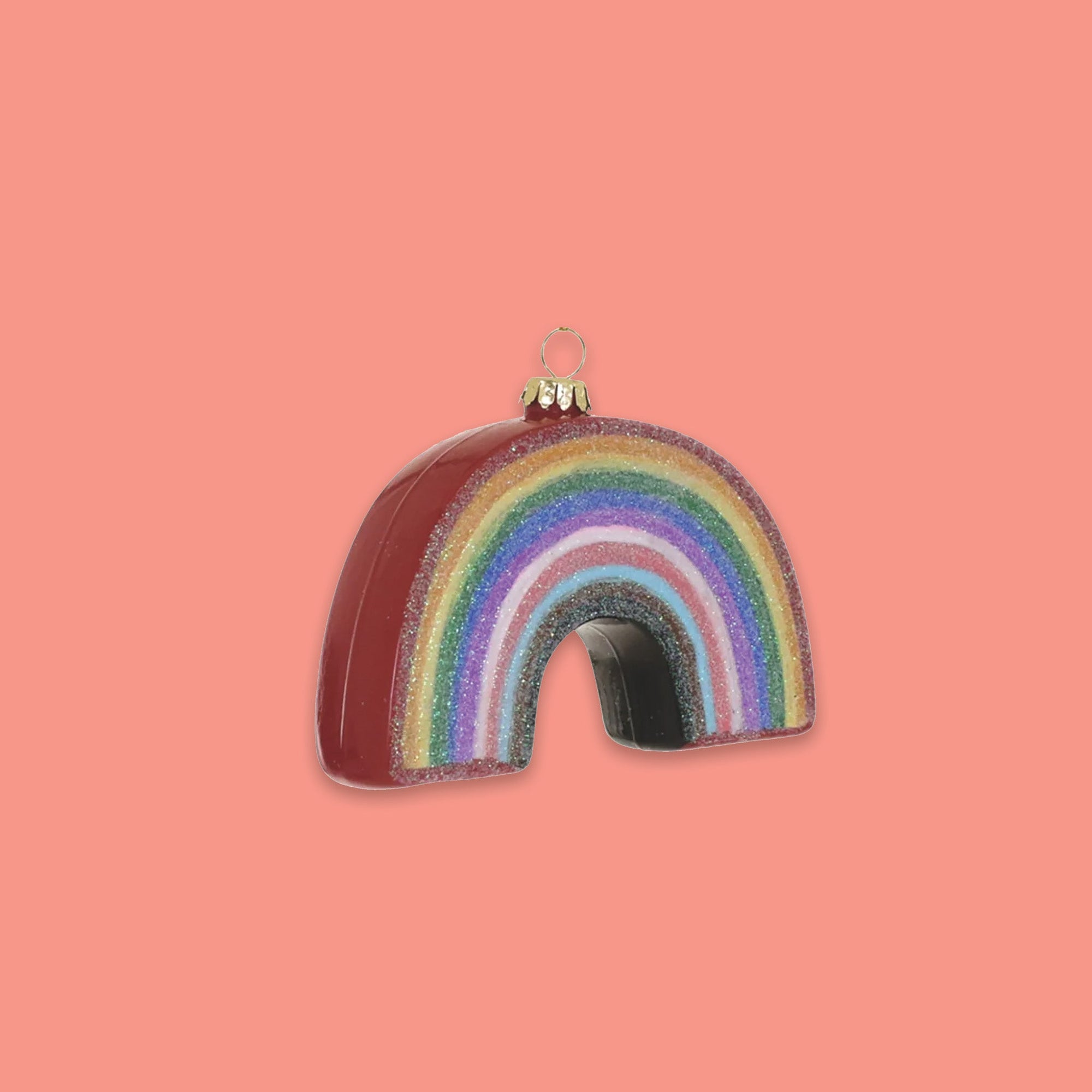 On a coral pink background sits an ornament. It is a glass rainbow and glitter colors of red, orange, yellow, green, blue, purple, pink, dark pink, and light blue.