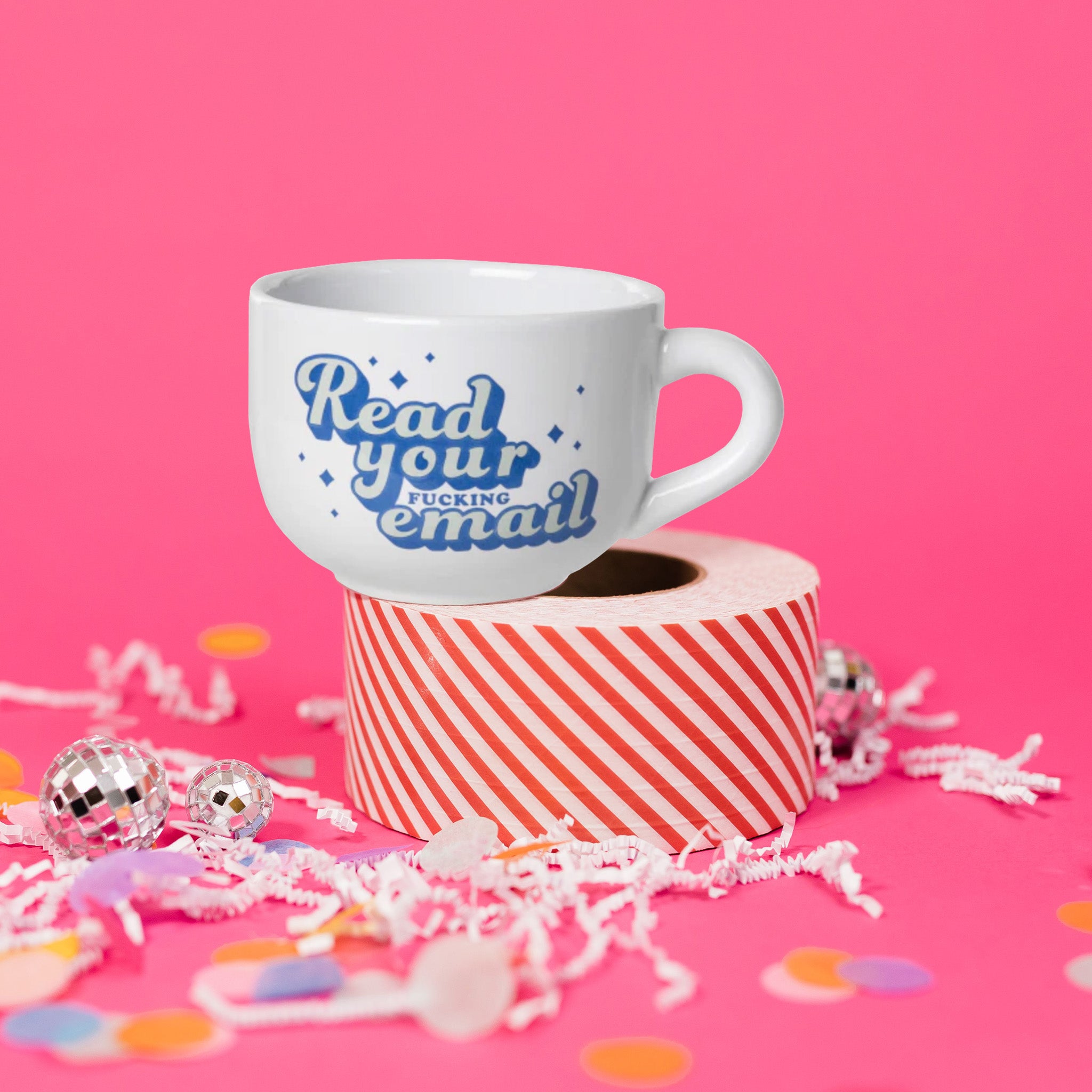 On a hot pink background sits a mug on top of a red and white striped packing tape with white crinkle and big, colorful confetti scattered around. There are mini disco balls. This white mug says "Read your fucking email" in a really cool retro lettering. It is in grey and blue and has blue stars scattered around the lettering.