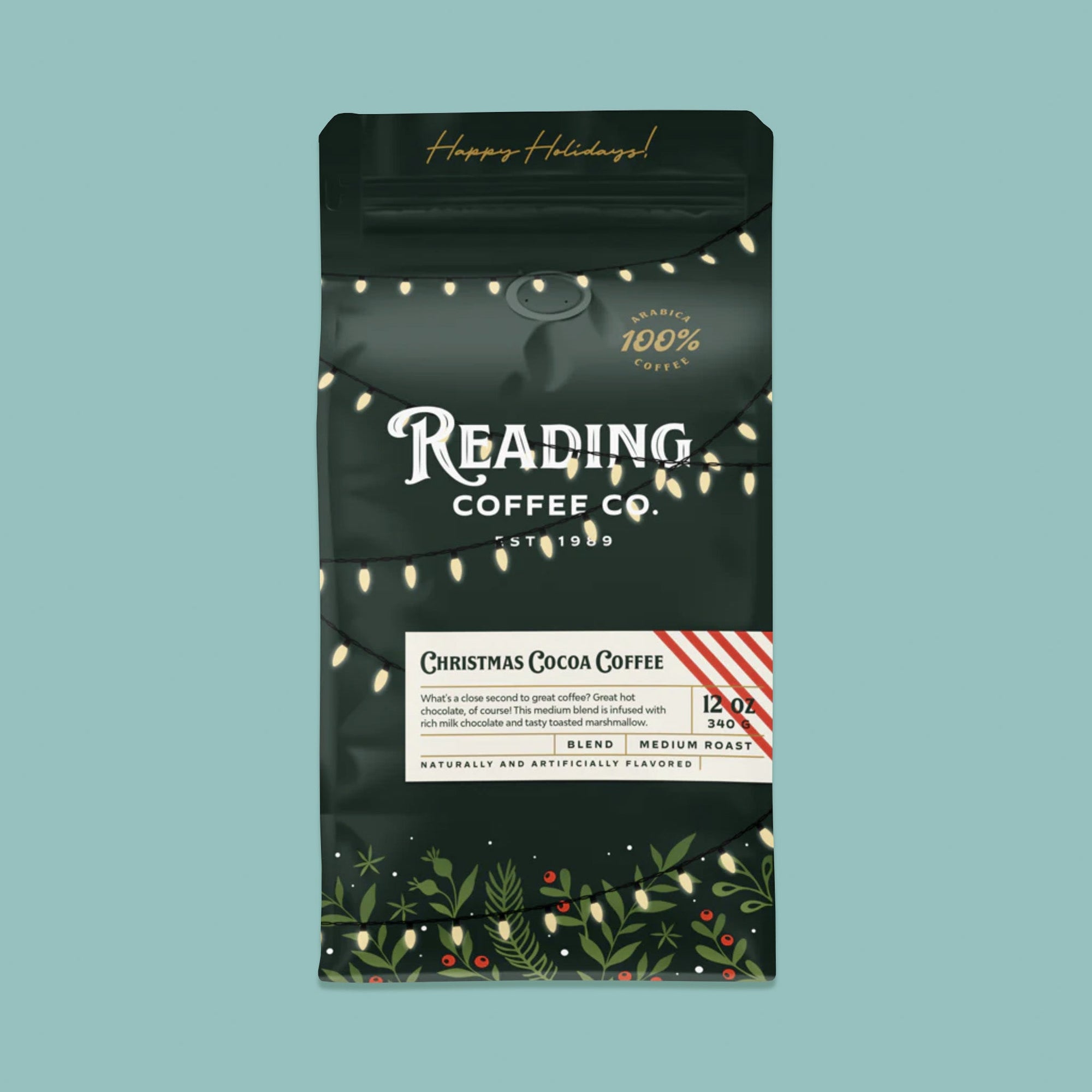 On a sage green background sits a dark green package of coffee. This package is gold, white, and dark green lettering. There are swags of lights and greenery with berries. It says "Happy Holidays!", "Reading Coffee Co. Est. 1989", "Christmas Cocoa Coffee Blend Medium Roast." It is '100% Arabica Coffee. It also says "What's a close second to great coffee? Great hot chocolate, of course! This medium blend is infused with rich milk chocolate and tasty toasted marshmallow." 12 oz 340g